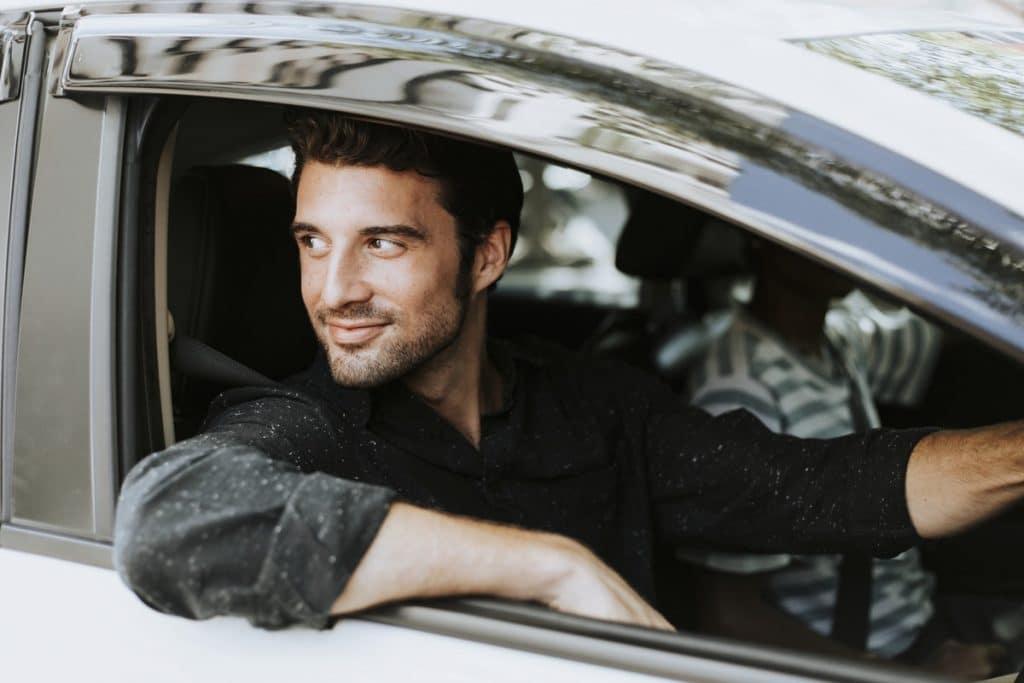 A man with dark hair, wearing a dark shirt, smiles while leaning out of the driver's side window of a white car, holding car keys. The interior of the car is partially visible, and another person is sitting in the passenger seat. Trees and buildings can be seen in the background.