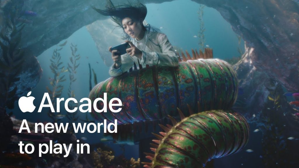 A person wearing a long coat is engrossed in playing a game on their phone while riding a large, colorful, caterpillar-like creature underwater. Text overlay reads "Apple Arcade: A new world to play in." The background features aquatic plants and rocky formations, perfect for a Super Bowl Ad.