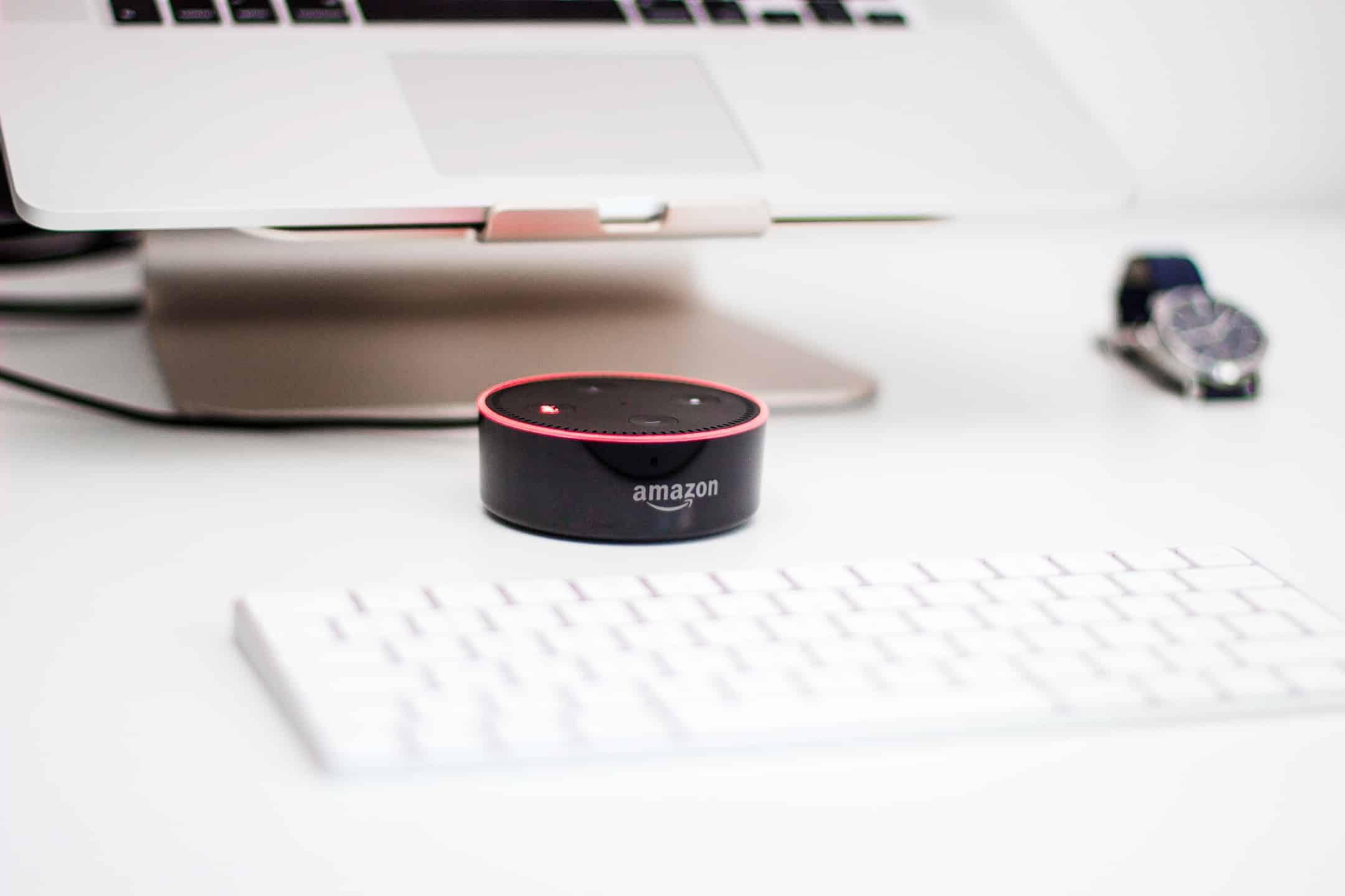 A black Amazon Echo Dot with a red ring light sits on a white desk in front of a laptop, which is on a stand. The keyboard is also on the desk, and an Apple wristwatch is placed near the laptop.