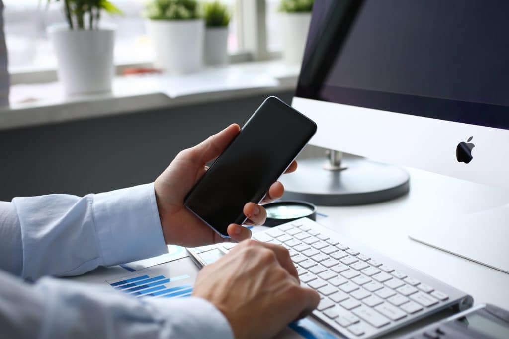 A person in a dress shirt holding a low-cost smartphone while using a desktop computer with an Apple logo. The desk has a keyboard, documents with blue charts, and potted plants near a window in the background.