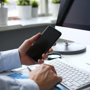 A person in a dress shirt holding a low-cost smartphone while using a desktop computer with an Apple logo. The desk has a keyboard, documents with blue charts, and potted plants near a window in the background.