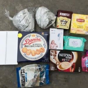 A collection of items arranged on the ground, including two wrapped bread loaves, Hershey's Kisses chocolates, Lipton tea, Danisa butter cookies, a pen, facial mask, Nescafé coffee sachets, wet wipes, and a bottle of hand soap—ideal for thoughtful care packages for employees in China.