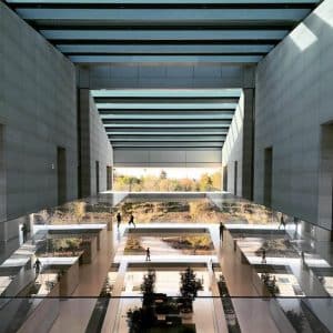 A modern, spacious atrium with multiple levels, featuring large glass windows and a reflective glass floor reminiscent of the Apple Headquarters. Trees and outdoor scenery are visible through the windows. Small figures of people are seen walking and using corridors in this case study-worthy space.