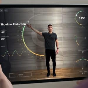 A person holds an iPad Pro displaying a fitness app that tracks shoulder abduction. The app shows a figure with one arm raised, with metrics including angles and ranges of motion. The background features a wooden floor and a wall with a circular graphic overlay, highlighting Apple's trackpad support for precise input.