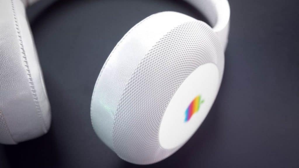 Close-up shot of white over-ear StudioPods with a circular outer casing. The earpiece features a small, colorful, retro-style rainbow stripe logo, set against a subtle mesh texture. The dark background contrasts with the clean Apple-inspired design of the headphones.