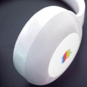 Close-up shot of white over-ear StudioPods with a circular outer casing. The earpiece features a small, colorful, retro-style rainbow stripe logo, set against a subtle mesh texture. The dark background contrasts with the clean Apple-inspired design of the headphones.