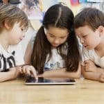 Three children are gathered around an Apple tablet on a wooden table. The girl in the middle is interacting with it while the two boys on either side watch intently. They are in a brightly lit room with colorful artwork and papers on the wall, showcasing elements of the education system.