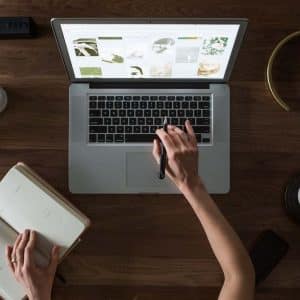 A person is seated at a wooden desk, working on a laptop and writing in an open notebook. The desk also has a plant, a cup of coffee, a smartphone, a candle, and a lamp. The laptop screen displays various images while the hand uses a stylus on the touchpad to recover deleted files.