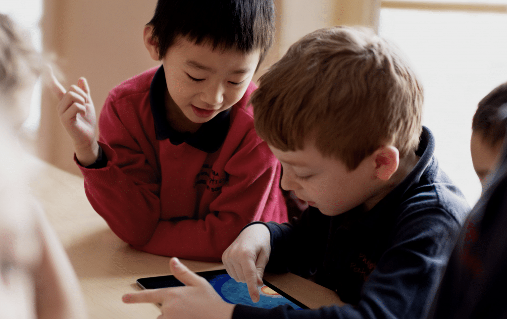 Two young boys engaged in a classroom setting, looking intently at an Apple tablet screen. The boy on the left is offering guidance by pointing at the screen and showing something to the boy on the right, who is interacting with it. They are surrounded by a warm, indoor environment supportive of remote learning.