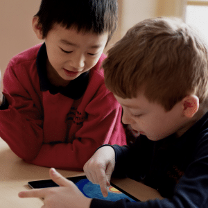 Two young boys engaged in a classroom setting, looking intently at an Apple tablet screen. The boy on the left is offering guidance by pointing at the screen and showing something to the boy on the right, who is interacting with it. They are surrounded by a warm, indoor environment supportive of remote learning.