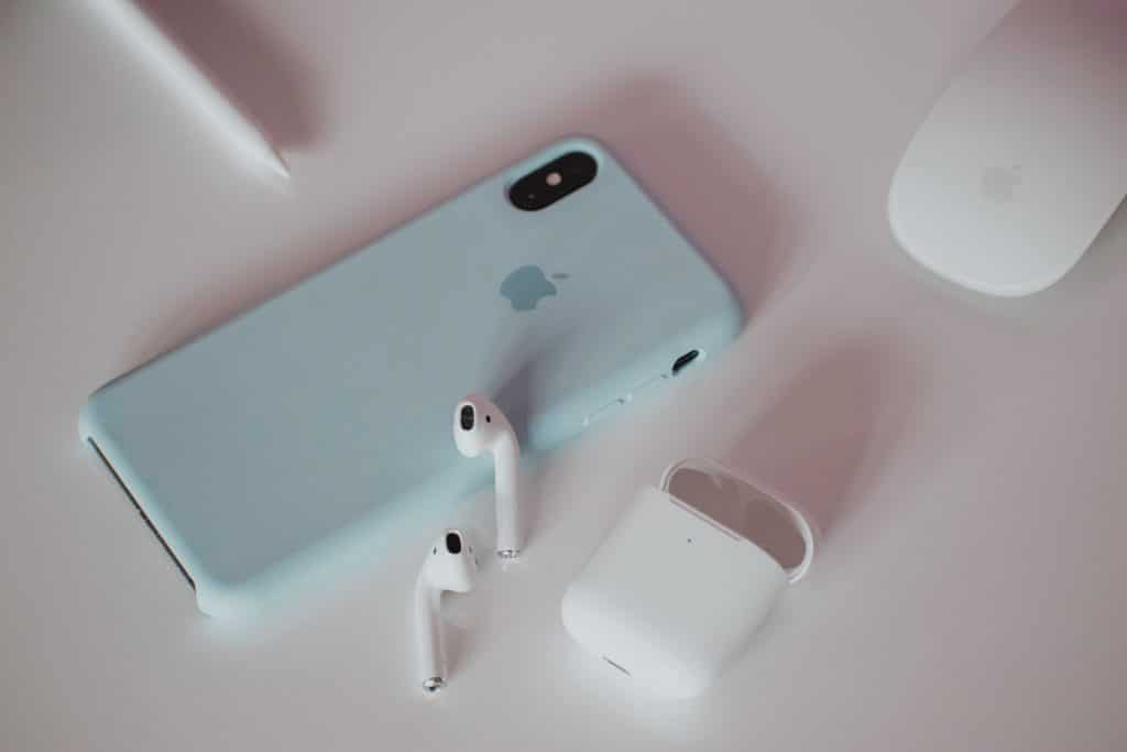 A smartphone with a light blue case lies on a white surface, accompanied by a pair of Apple AirPods in their white charging case. Nearby, a computer mouse and stylus add to the tech-themed setup, reminiscent of 2020's sleek gadget aesthetic.