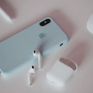 A smartphone with a light blue case lies on a white surface, accompanied by a pair of Apple AirPods in their white charging case. Nearby, a computer mouse and stylus add to the tech-themed setup, reminiscent of 2020's sleek gadget aesthetic.
