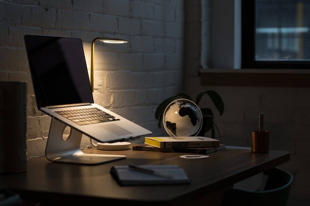 A dimly lit workspace with a laptop on a stand, possibly undergoing important tasks for Mac users, a small lamp casting warm light, a globe, and several books on the desk. A notebook and pen are also on the desk, and there's a potted plant in the background near a window.