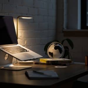 A dimly lit workspace with a laptop on a stand, possibly undergoing important tasks for Mac users, a small lamp casting warm light, a globe, and several books on the desk. A notebook and pen are also on the desk, and there's a potted plant in the background near a window.