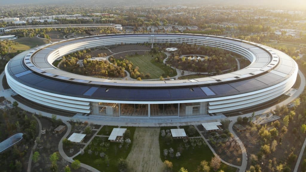 An aerial view of the large, circular Apple Park Campus showcases its futuristic design and a central garden area surrounded by the ring structure. The surrounding landscape is lush with trees and greenery, captured beautifully in drone footage as the sun sets in the background.