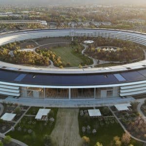 An aerial view of the large, circular Apple Park Campus showcases its futuristic design and a central garden area surrounded by the ring structure. The surrounding landscape is lush with trees and greenery, captured beautifully in drone footage as the sun sets in the background.