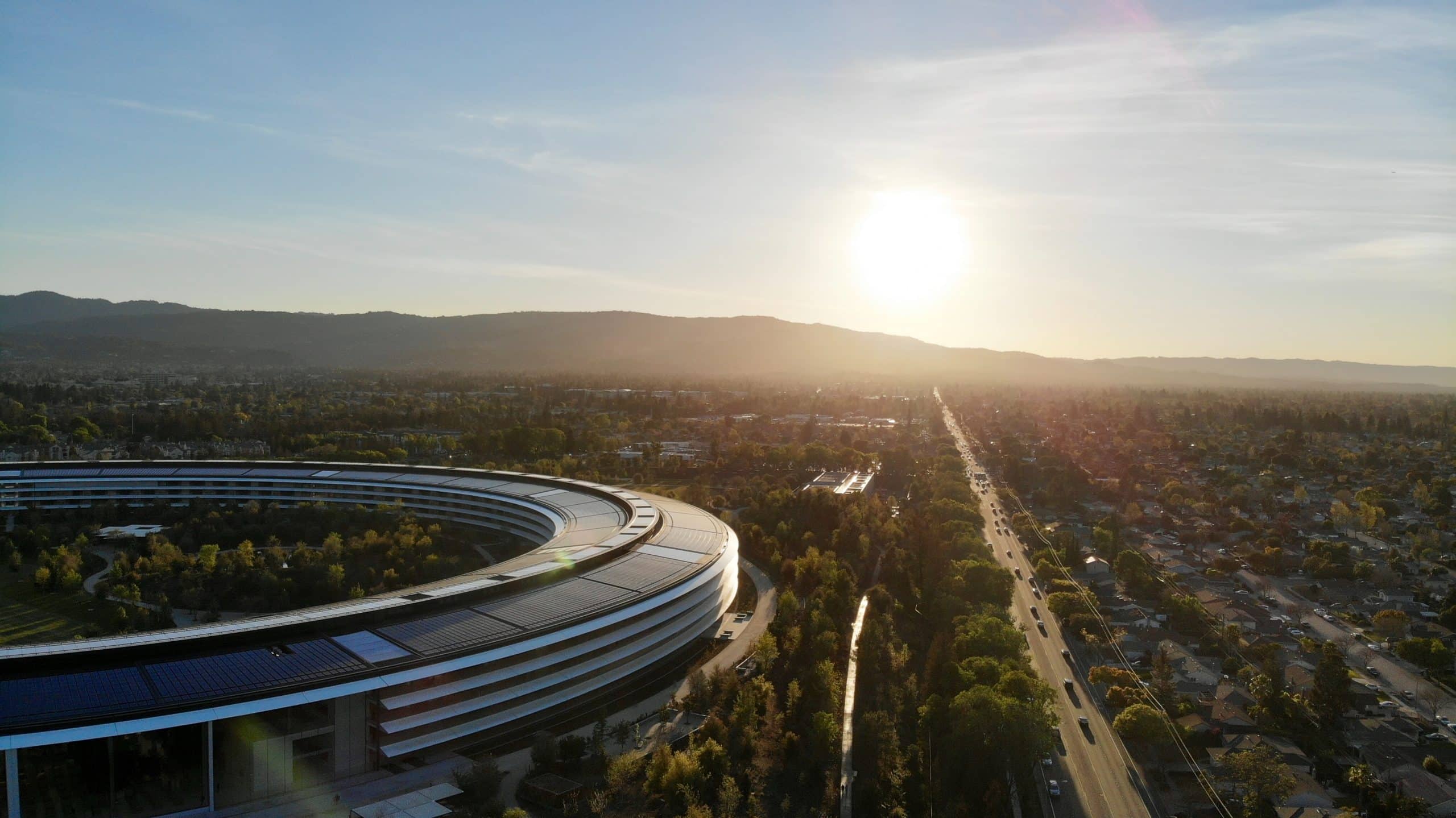 Aerial view of a large, circular building with solar panels on its roof, surrounded by trees and adjacent to a roadway. The sun is setting in the background, casting a warm glow over the distant mountains and suburban landscape, hinting at the tranquility that belies the aftermath of the March event.