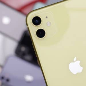 Close-up of a yellow Apple iPhone 12 being held in hand, showcasing its dual camera setup and Apple logo on the back. In the blurred background, various iPhone models in different colors are laid out, reminding us of the delayed releases during the coronavirus outbreak.