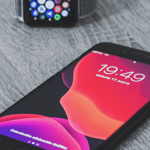 A smartphone displaying the time "19:49" and the date in a foreign language is lying on a wooden surface. In the background, an Apple Watch is visible, showcasing Apple's signature home screen design with various app icons.