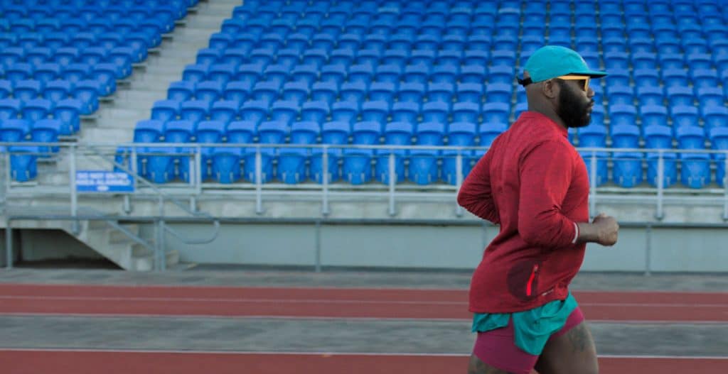 A person in a red long-sleeve shirt, green shorts, and a green and yellow cap is jogging on a track in front of empty blue stadium seats, using their fitness app on an Apple device running iOS 14.