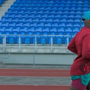 A person in a red long-sleeve shirt, green shorts, and a green and yellow cap is jogging on a track in front of empty blue stadium seats, using their fitness app on an Apple device running iOS 14.