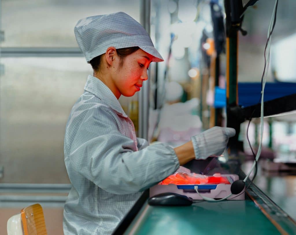 A worker wearing a white cap and lab coat operates machinery at an assembly line in a manufacturing facility. During the second quarter of 2020, they are handling iPhone production components with precision under bright lighting, emphasizing focus and concentration in an industrial setting.