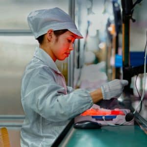 A worker wearing a white cap and lab coat operates machinery at an assembly line in a manufacturing facility. During the second quarter of 2020, they are handling iPhone production components with precision under bright lighting, emphasizing focus and concentration in an industrial setting.