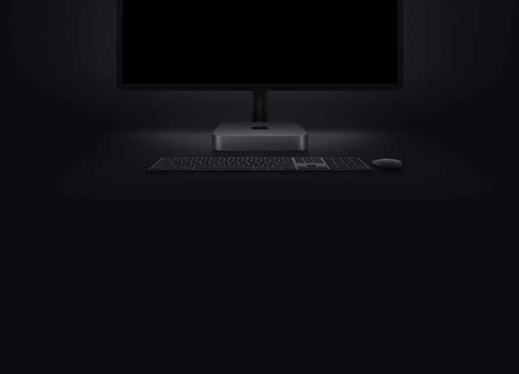 A dark-themed workspace setup showcasing a sleek monitor, the latest Mac mini with a storage bump, a wireless keyboard, and a mouse, all placed against a dark background. The devices have a minimalist design, emphasizing a modern and clean aesthetic.