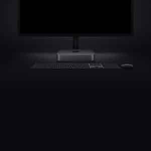 A dark-themed workspace setup showcasing a sleek monitor, the latest Mac mini with a storage bump, a wireless keyboard, and a mouse, all placed against a dark background. The devices have a minimalist design, emphasizing a modern and clean aesthetic.