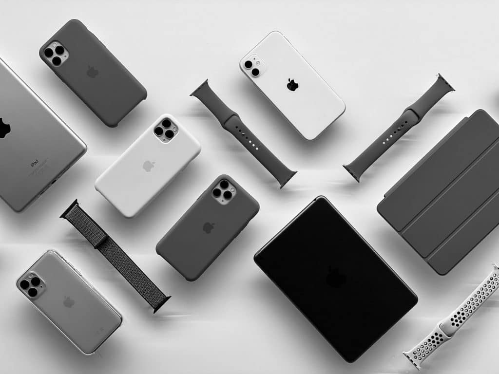 A flat lay of various Apple products in grayscale, including iPhones, an iPad, an Apple Watch with different bands, and a MacBook. The items are arranged neatly against a white background, highlighting the sleek design amid COVID-19 related shortages.