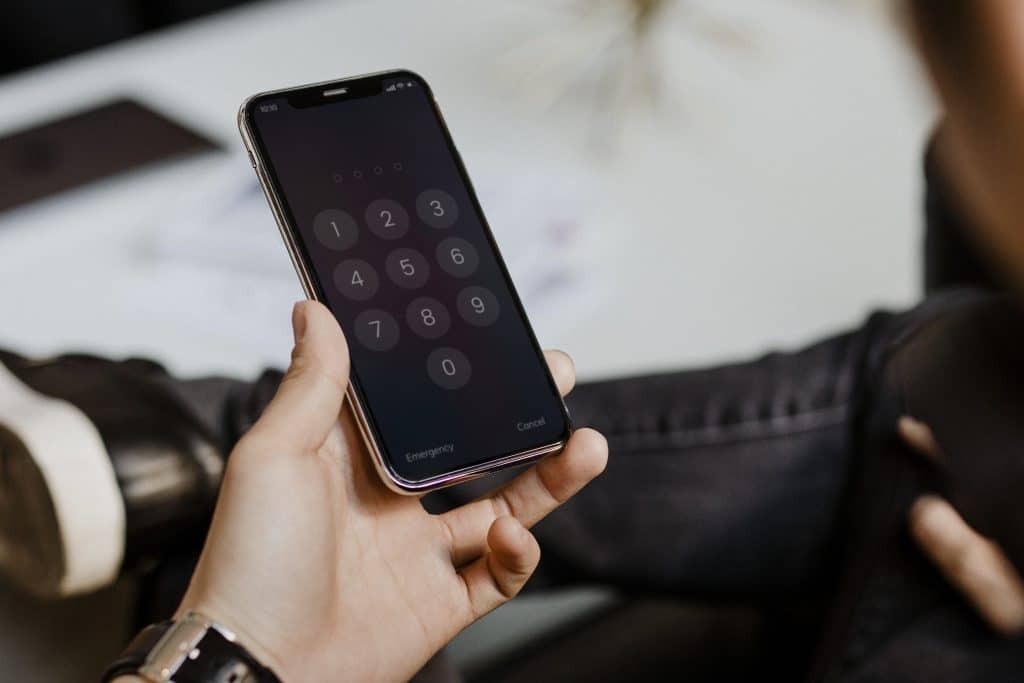 A person holding a smartphone with a passcode entry screen, displaying numbers from 0 to 9 in a keypad format. The person's other hand is relaxed on their lap, and they are dressed in casual clothing. The background is slightly blurred, emphasizing the importance of iPhone security tips for a secure iPhone.