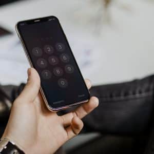 A person holding a smartphone with a passcode entry screen, displaying numbers from 0 to 9 in a keypad format. The person's other hand is relaxed on their lap, and they are dressed in casual clothing. The background is slightly blurred, emphasizing the importance of iPhone security tips for a secure iPhone.