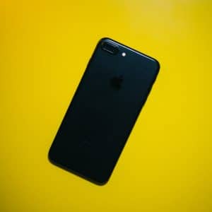 A black iPhone 7 lies face down on a bright yellow surface, showcasing the Apple logo and dual camera on the back.