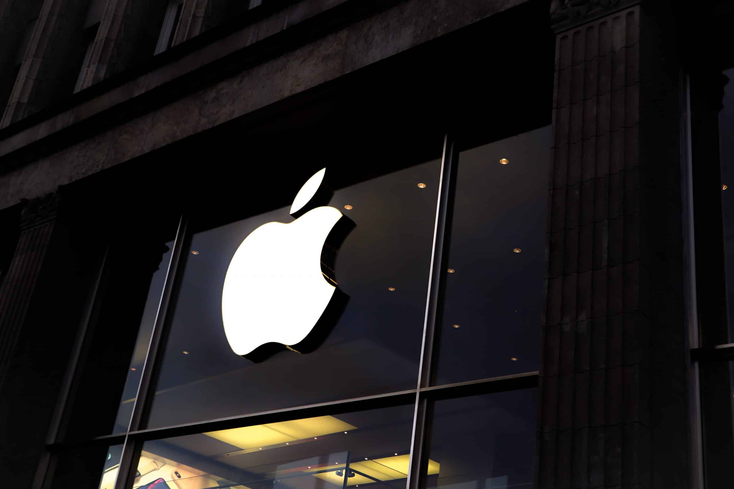 An illuminated Apple logo is prominently displayed on the glass of an Apple Store's exterior, pausing passersby with its glow. The store's interior lights are visible, and the building's modern architectural details surround the logo. The night sky is reflected in the windows, hinting at a magical evening ahead for Today at Apple.