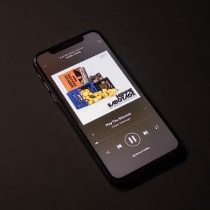 A smartphone running iOS 14 lies on a dark surface, displaying Spotify with the song "Pay For The Silence" by Hippie Sabotage. The album cover shows crates filled with fruits, including apples. Music controls are visible at the bottom of the screen.