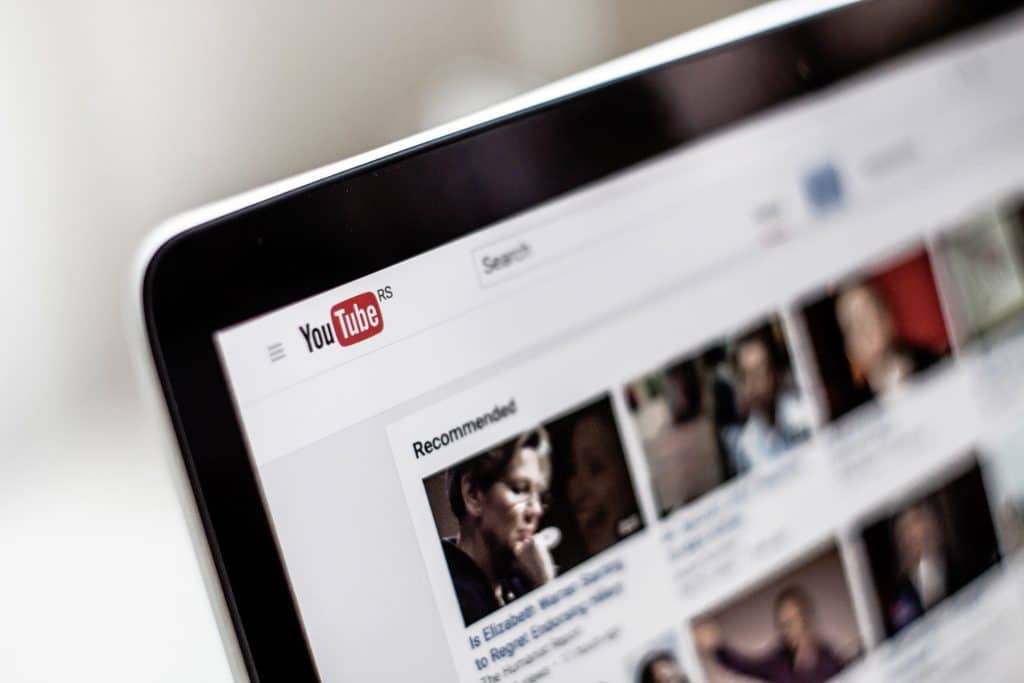 Close-up of a computer screen showing the YouTube homepage with the search bar at the top and several video thumbnails in the 'Recommended' section below. The red and white YouTube logo is prominently displayed in the upper left corner, highlighting popular videos on coronavirus and tips for improving streaming quality.