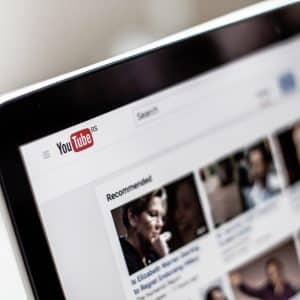 Close-up of a computer screen showing the YouTube homepage with the search bar at the top and several video thumbnails in the 'Recommended' section below. The red and white YouTube logo is prominently displayed in the upper left corner, highlighting popular videos on coronavirus and tips for improving streaming quality.