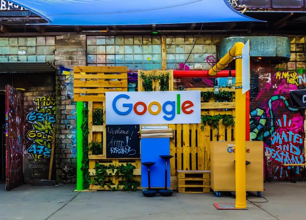 A colorful, outdoor setup with a "Google" sign displayed on a wooden pallet backdrop, reminiscent of the Google I/O conference. The area is decorated with graffiti and plants, and is sheltered by an overhead canopy. Various furniture pieces, including chairs and stands, are also present despite coronavirus fears.