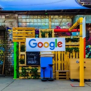 A colorful, outdoor setup with a "Google" sign displayed on a wooden pallet backdrop, reminiscent of the Google I/O conference. The area is decorated with graffiti and plants, and is sheltered by an overhead canopy. Various furniture pieces, including chairs and stands, are also present despite coronavirus fears.