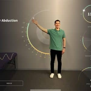 A man in a green polo shirt performs a shoulder abduction exercise in front of a digital display, which showcases metrics like range goal (180°), current range (131°), active range (113°), shoulder alignment (1°), and spinal alignment (0°). Using ARKit 3.5 on the latest iOS updates, an Apple device tracks his progress. A bench is visible