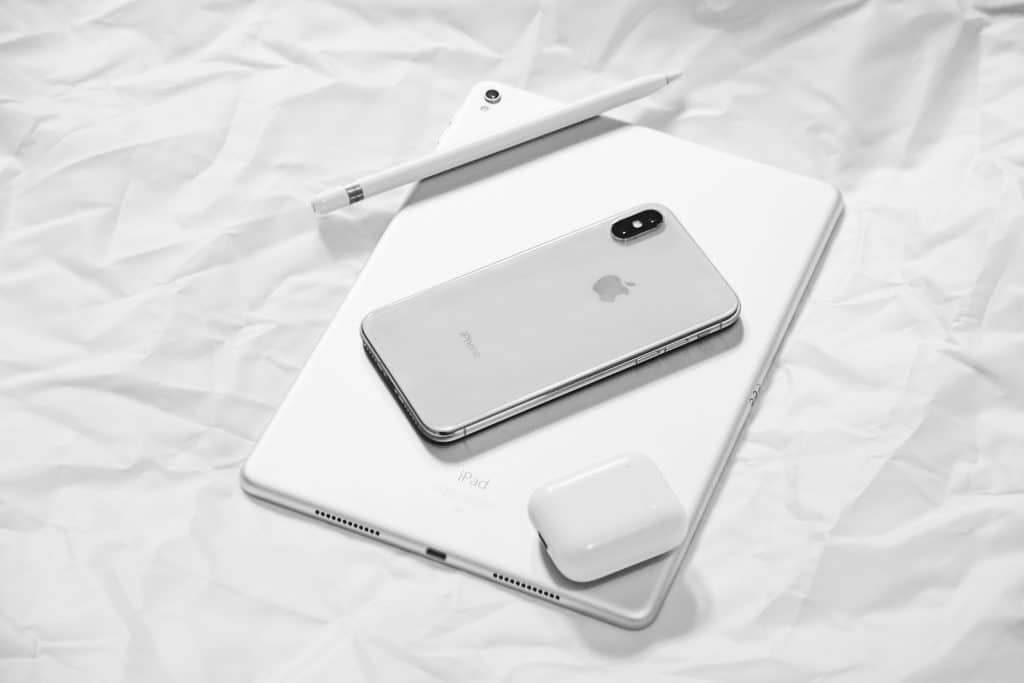 A silver iPhone, white Apple Pencil, white iPad, and white AirPods charging case rest on crumpled white paper. The sleek, modern design of the Apple devices is highlighted in the minimalist black-and-white composition—a perfect visual for anyone considering switching to Apple.