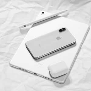 A silver iPhone, white Apple Pencil, white iPad, and white AirPods charging case rest on crumpled white paper. The sleek, modern design of the Apple devices is highlighted in the minimalist black-and-white composition—a perfect visual for anyone considering switching to Apple.