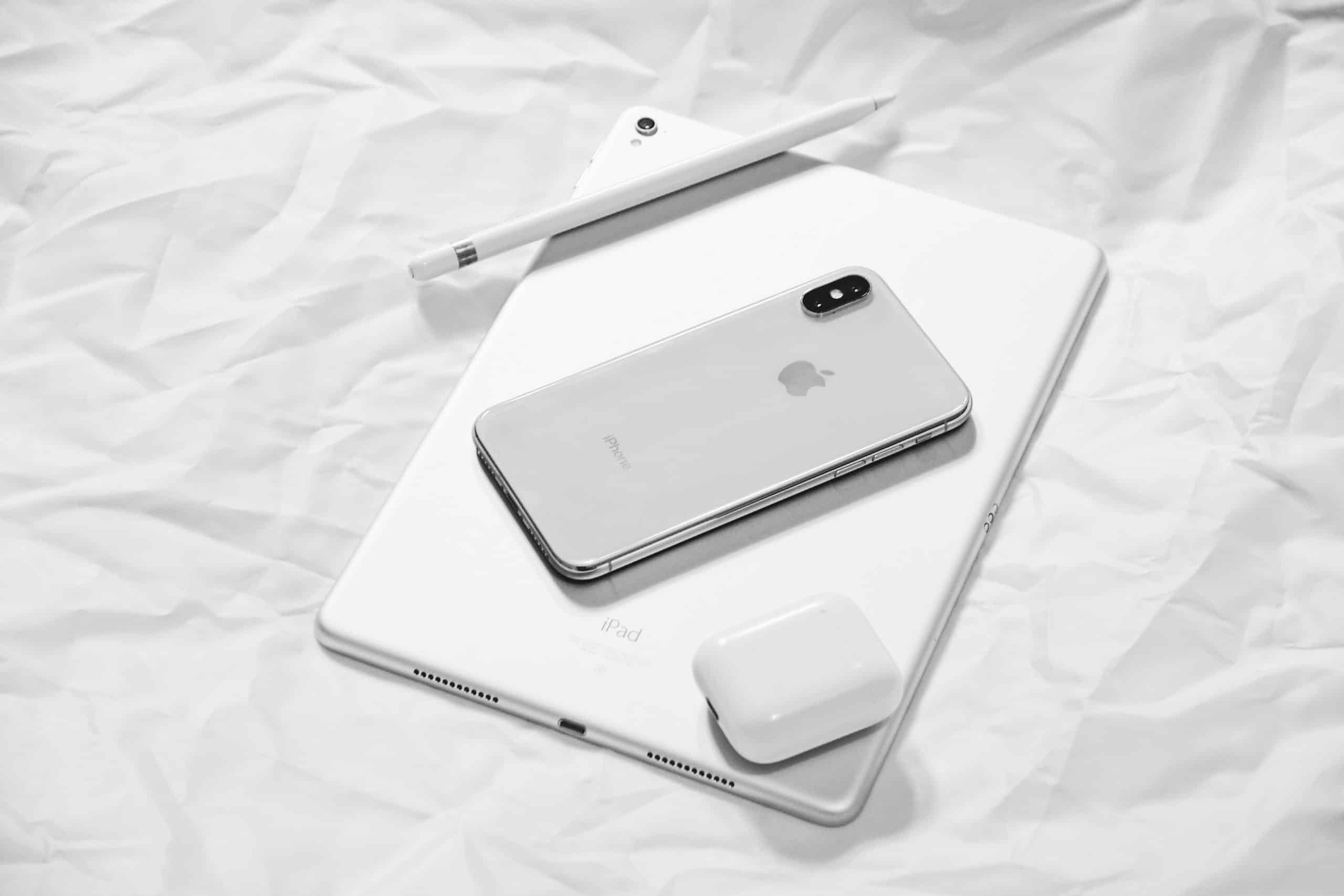 A silver iPhone, white Apple Pencil, white iPad, and white AirPods charging case rest on crumpled white paper. The sleek, modern design of the Apple devices is highlighted in the minimalist black-and-white composition—a perfect visual for anyone considering switching to Apple.