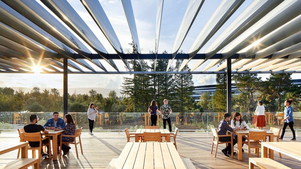 Outdoor seating area with wooden tables and chairs under a modern pergola structure with an open slatted roof. Office-based employees are engaged in conversation or working on laptops, enjoying the sunlight streaming through the slats. Tall trees and a building are visible in the distance.