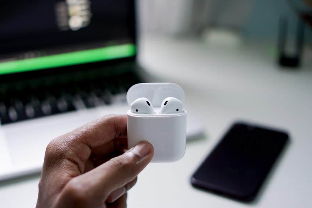 A person holds an open white case containing a pair of wireless earbuds. In the background, there is a blurred view of a laptop and a smartphone placed on a white table, perfect for exploring AirPods tips.