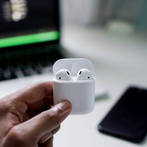 A person holds an open white case containing a pair of wireless earbuds. In the background, there is a blurred view of a laptop and a smartphone placed on a white table, perfect for exploring AirPods tips.