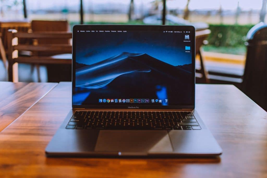 A MacBook Pro with a dark desert landscape wallpaper on its screen sits on a wooden table in a cafe. The background shows blurred tables, chairs, and large windows letting in daylight. The Mac owner seems ready to perform various tasks, as the desktop displays several icons and application shortcuts.