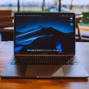 A MacBook Pro with a dark desert landscape wallpaper on its screen sits on a wooden table in a cafe. The background shows blurred tables, chairs, and large windows letting in daylight. The Mac owner seems ready to perform various tasks, as the desktop displays several icons and application shortcuts.