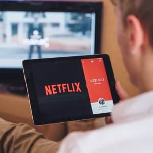 A person holding a tablet displaying the Netflix logo while seated on a couch. In the background, a television shows a blurred screen, indicating a program is playing. The person appears to be setting up or browsing Netflix on the tablet, likely checking streaming quality during coronavirus lockdowns.