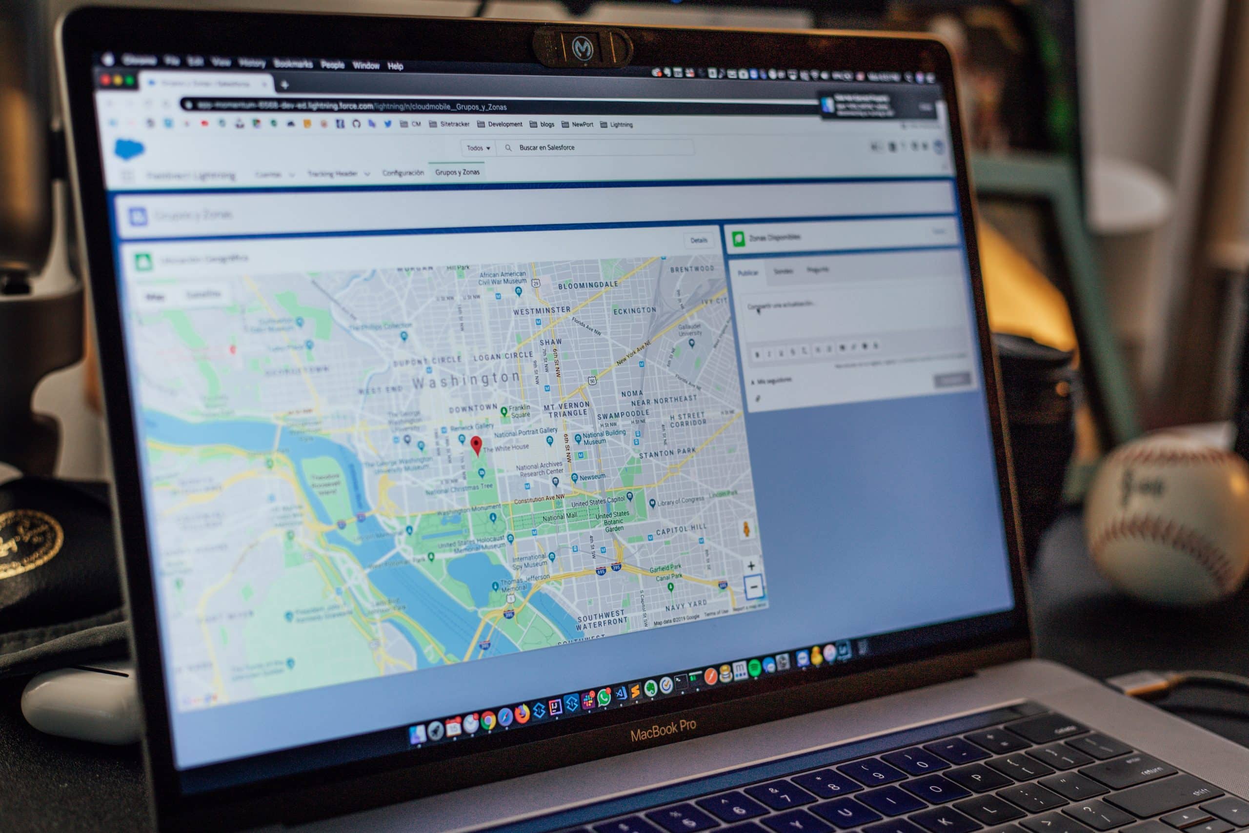 A MacBook Pro laptop screen displays a map of Washington, D.C., with a red location pin marking a specific area. Various desktop objects, including a baseball, are partially visible in the background. The browser window shows navigation and search options, possibly for US officials or tech giants.